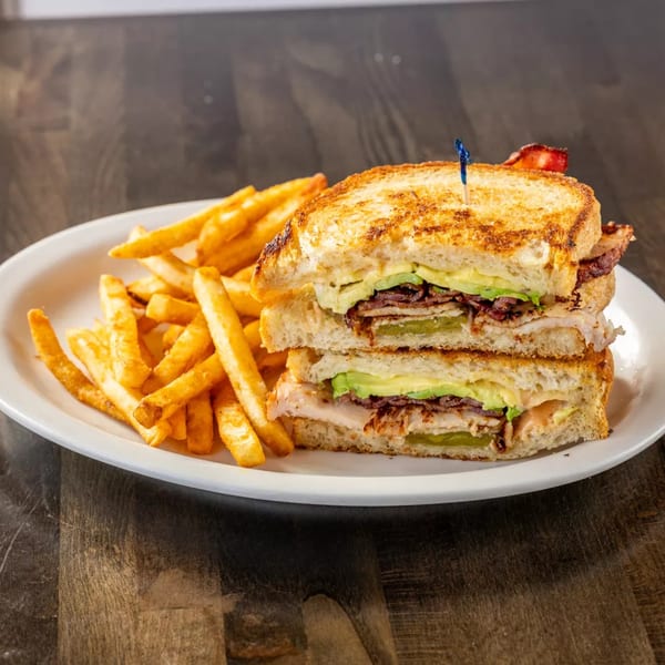 Grilled sandwich with bacon, avocado, and pickles, served with a side of golden french fries.