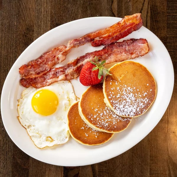 Breakfast plate with crispy bacon, sunny-side-up egg, pancakes with powdered sugar, and a strawberry.