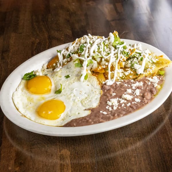 A white oval plate holds chilaquiles with two fried eggs and refried beans on a wooden table.