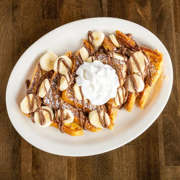 Golden french toast topped with banana slices, chocolate drizzle, powdered sugar, and whipped cream.